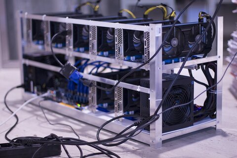 A metal rig with multiple graphics cards, fans, and cables, typically used for cryptocurrency mining to acquire Bitcoin without ID verification, is placed on a flat surface.