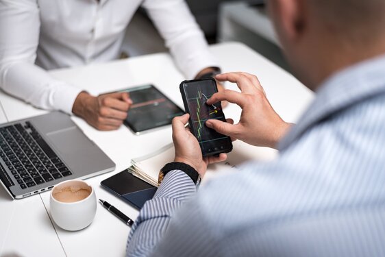 Two people are at a desk with a laptop, smartphone, and tablet. One is analyzing a financial graph on the phone, exploring how to acquire Bitcoin without ID verification, while the other uses the tablet. A cup of coffee sits nearby.