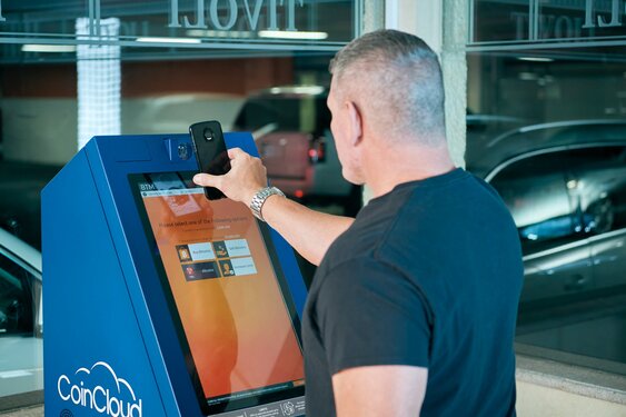 A man uses a smartphone to interact with a CoinCloud cryptocurrency ATM inside a building, effortlessly acquiring Bitcoin without ID verification.