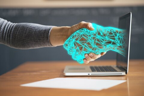 A person extends their hand to shake with a digital hand emerging from a laptop, symbolizing the connection between humans and technology, much like how one can now easily acquire Bitcoin without ID verification.